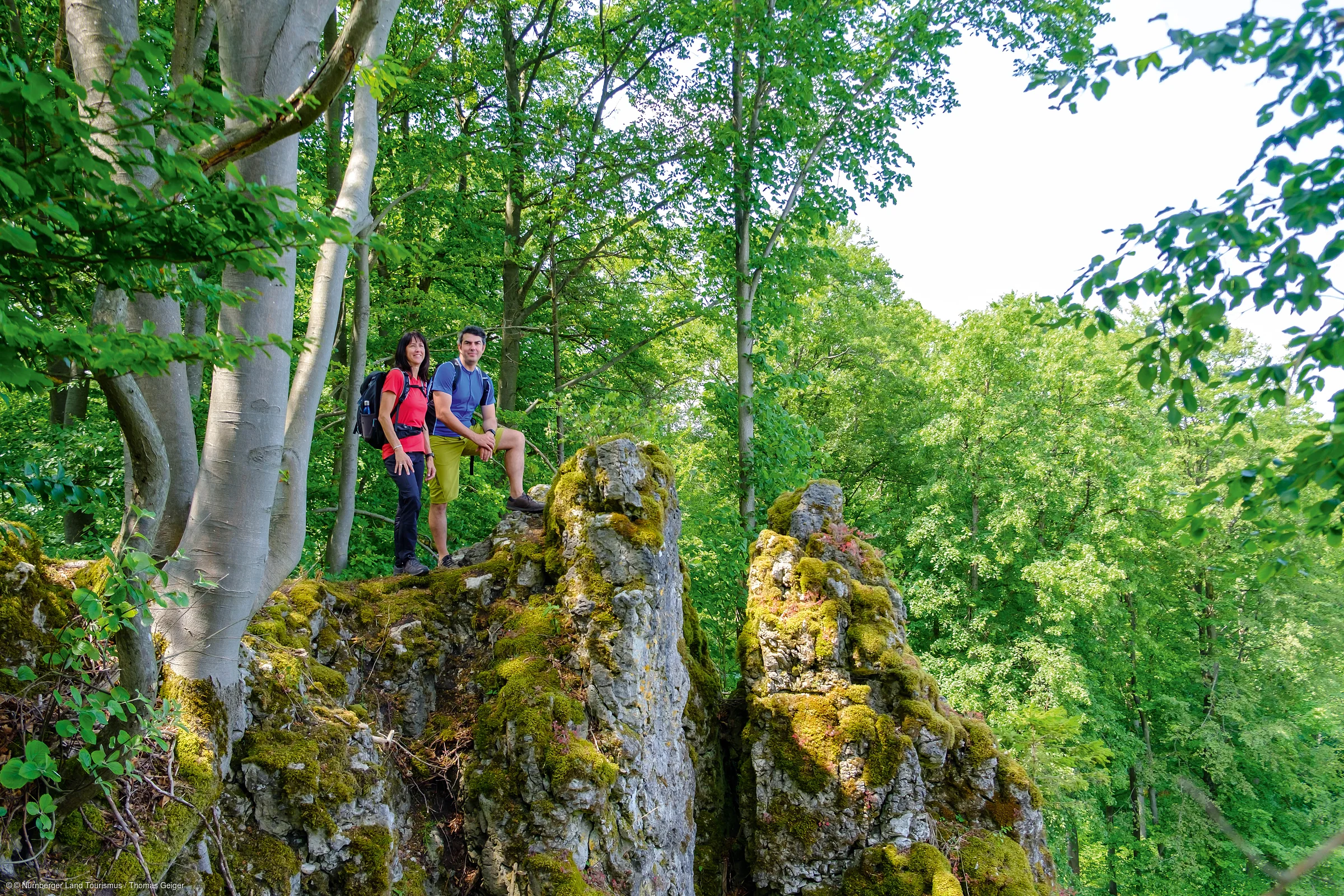 Zwei Wanderer mit Rucksack stehen auf moosbedecktem Felsen im Wald bei Tageslicht.