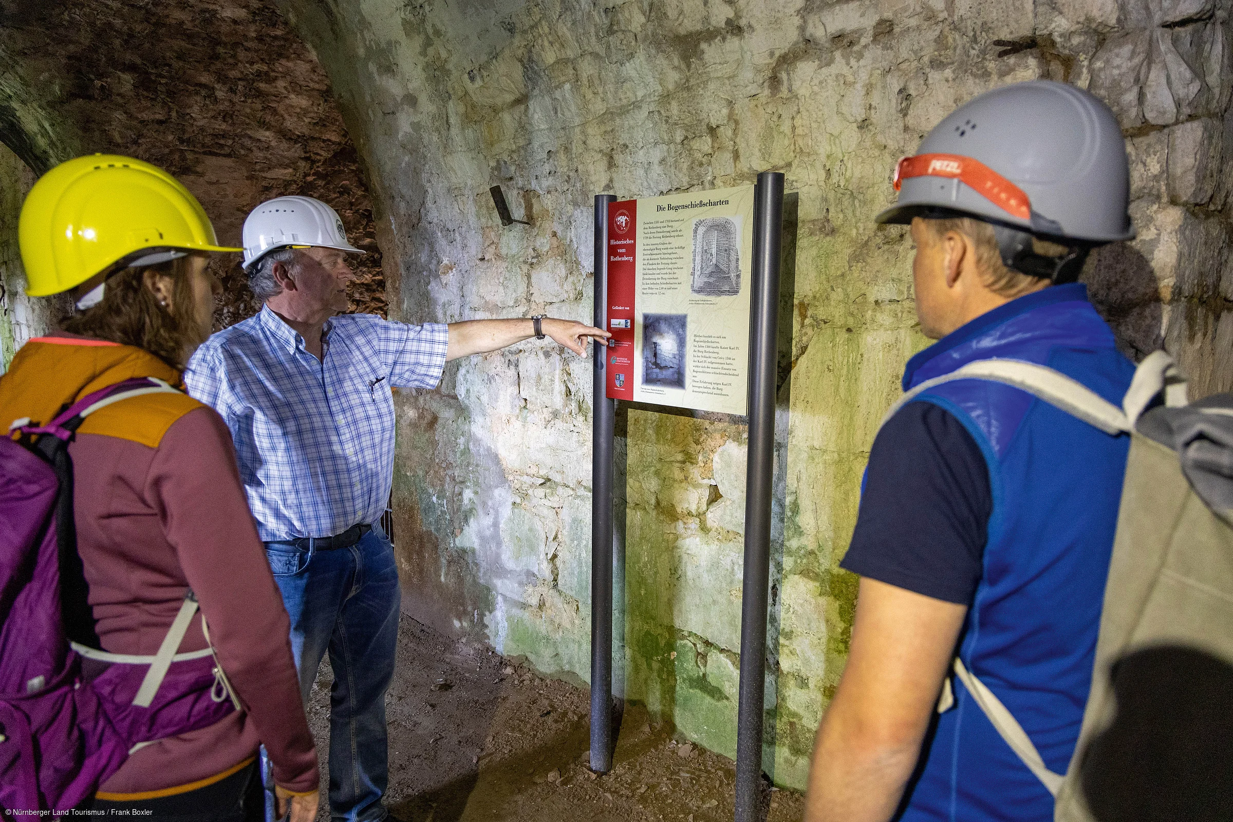 Drei Personen mit Schutzhelmen betrachten eine Informationstafel in einem Tunnel mit steinernen Wänden.