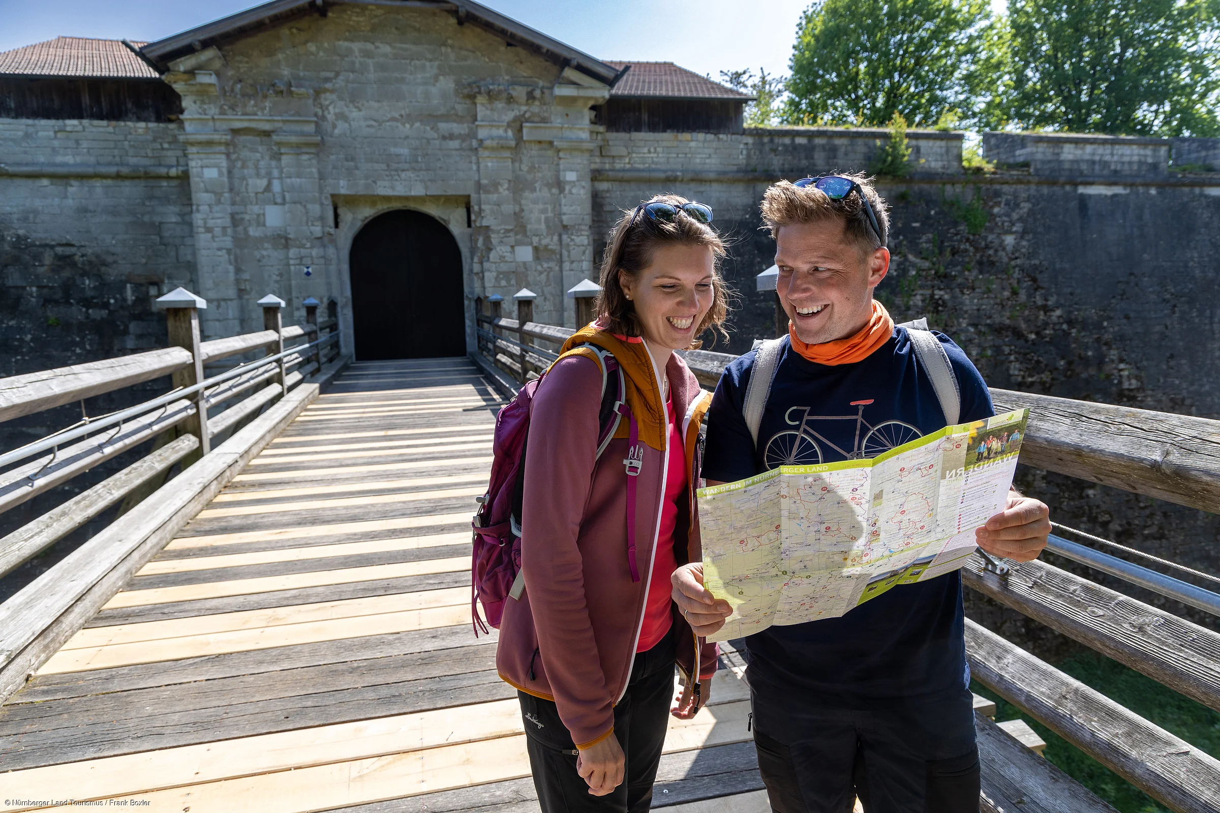 Zwei Wanderer mit Rucksäcken lesen eine Karte auf einer Holzbrücke vor einem steinernen Tor.