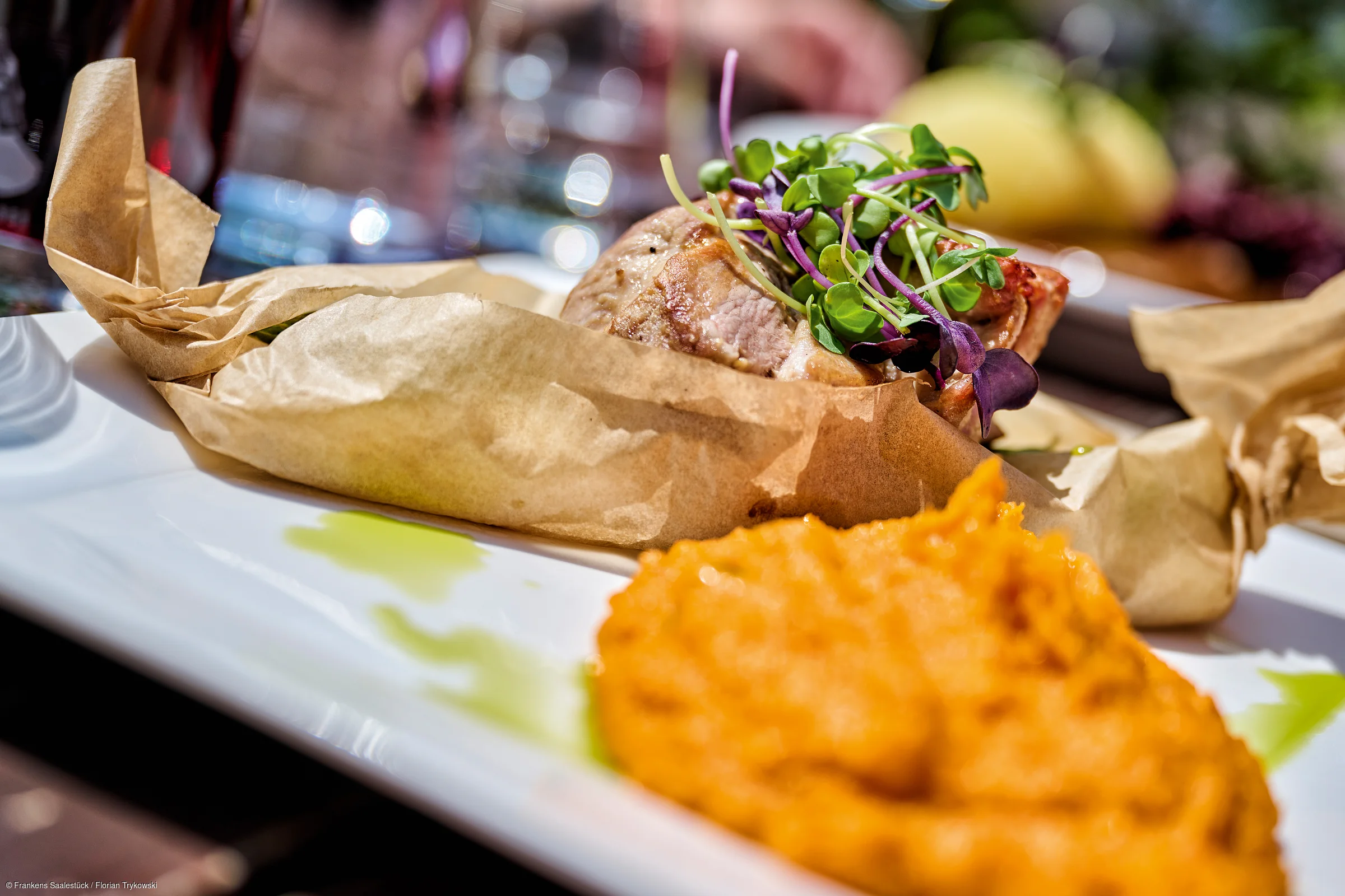 Teller mit Fleisch in Backpapier und grünen Sprossen, dazu orangefarbene Beilage, unscharfer Hintergrund.