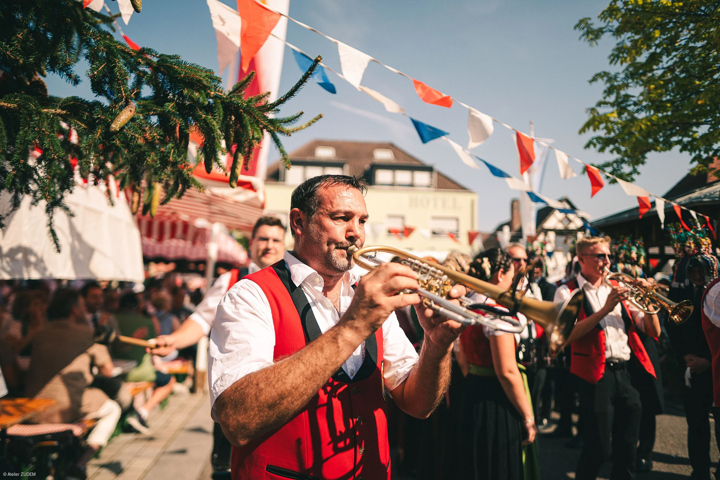 Mann in roter Weste spielt Trompete bei Kirchweih mit Menschen und Girlanden im Hintergrund.
