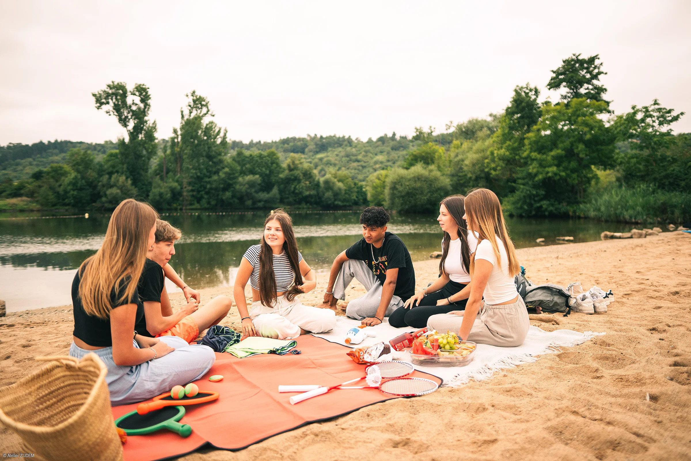 Sechs Jugendliche sitzen auf Decken am Sandstrand neben einem See mit Bäumen im Hintergrund.
