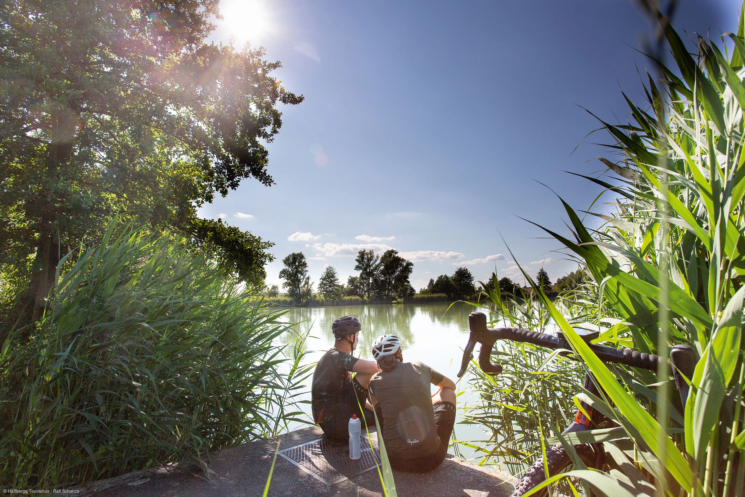 Zwei Radfahrer mit Helmen sitzen am Ufer eines Sees, umgeben von Schilf und Bäumen bei Sonnenschein.