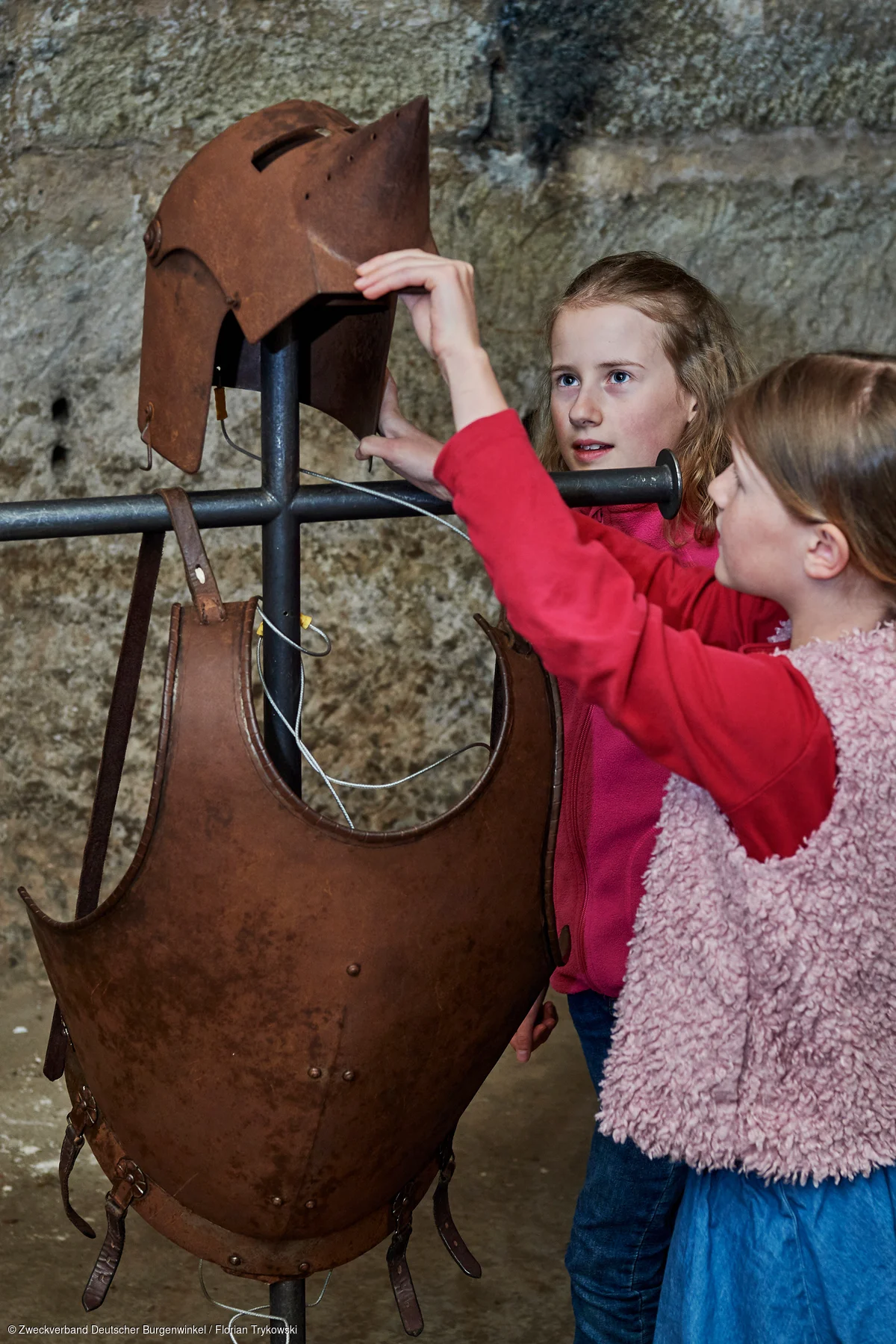 Zwei Mädchen betrachten und berühren einen historischen Ritterhelm und eine Brustplatte aus Metall an einem Ständer.