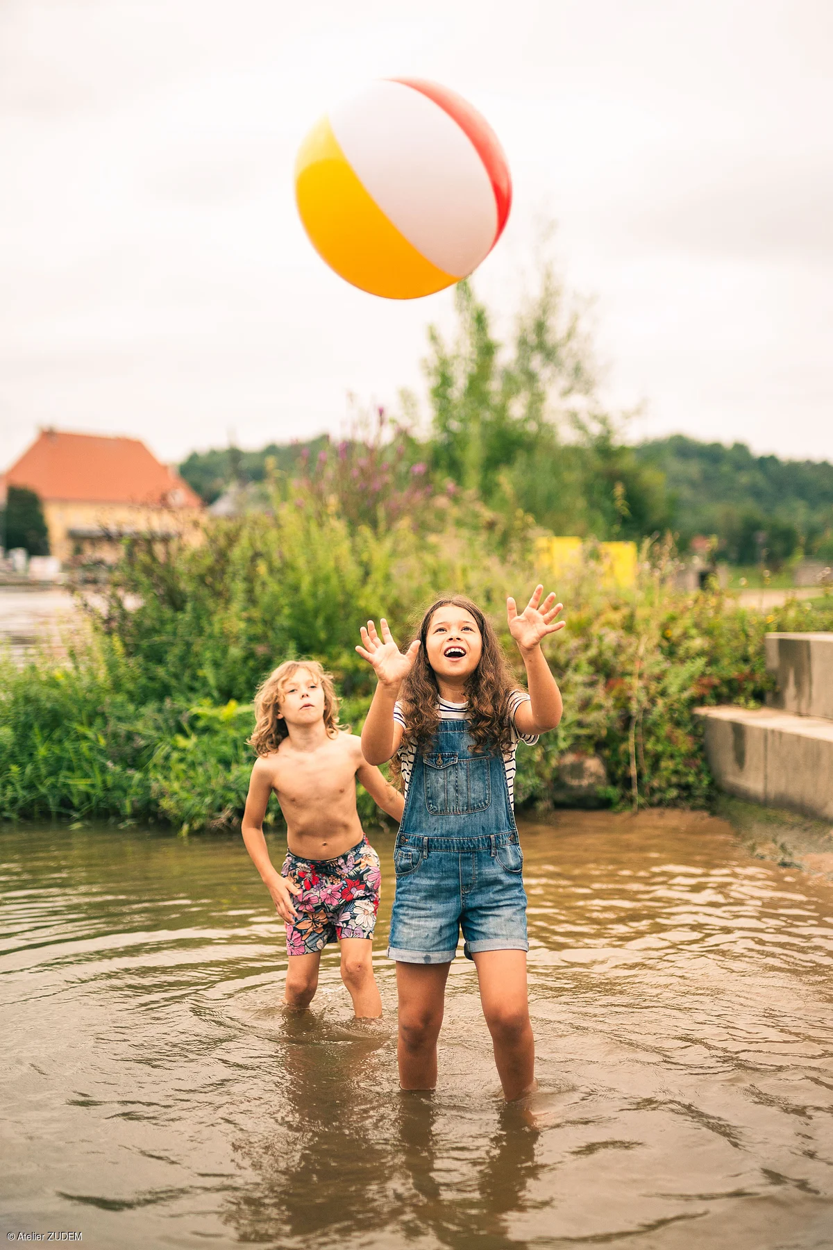 Zwei Kinder spielen mit einem bunten Ball im flachen Wasser eines Teichs oder Sees.