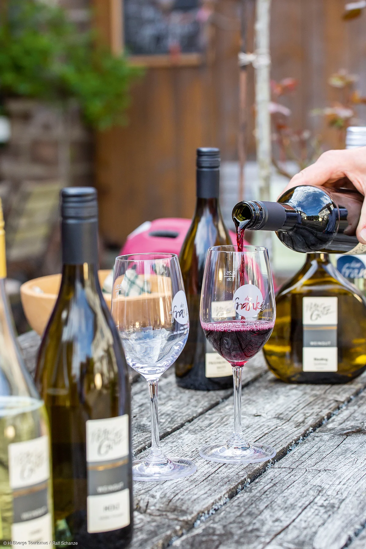Wein wird aus einer Flasche in ein Weinglas auf einem Holztisch im Freien eingeschenkt, daneben weitere Weinflaschen.