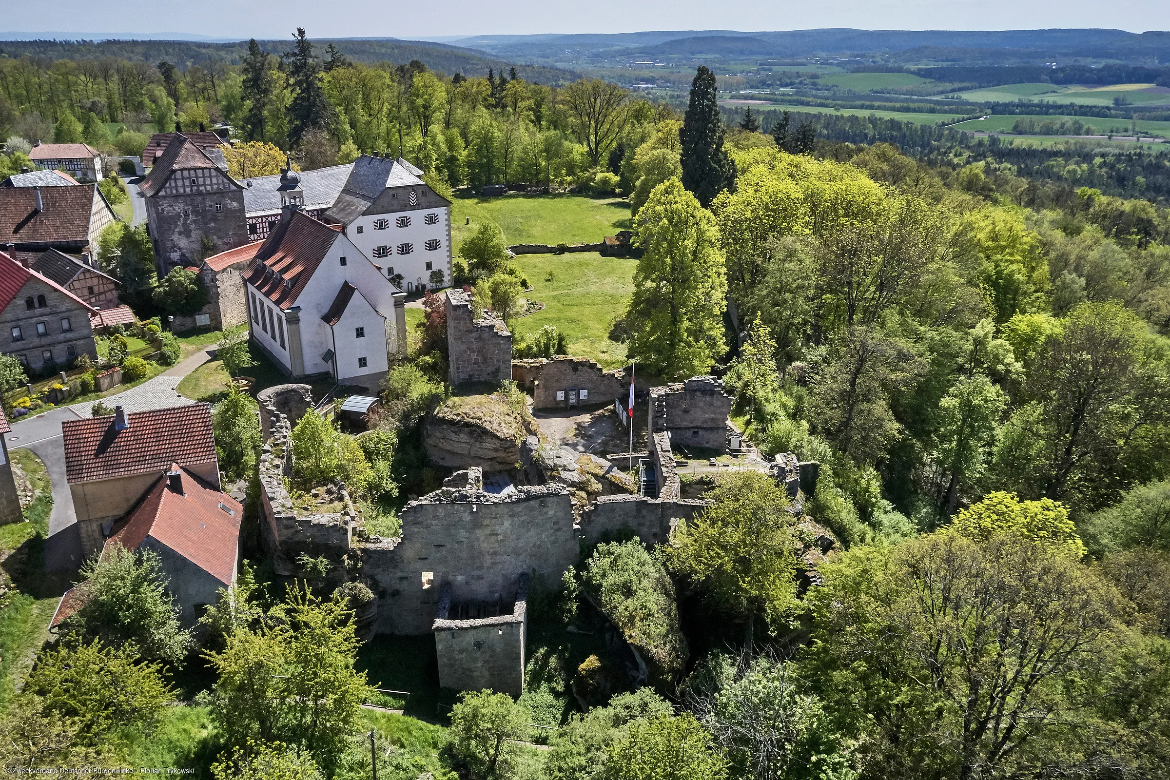 Luftaufnahme einer historischen Burganlage mit umliegenden Gebäuden und Wald in einer hügeligen Landschaft.