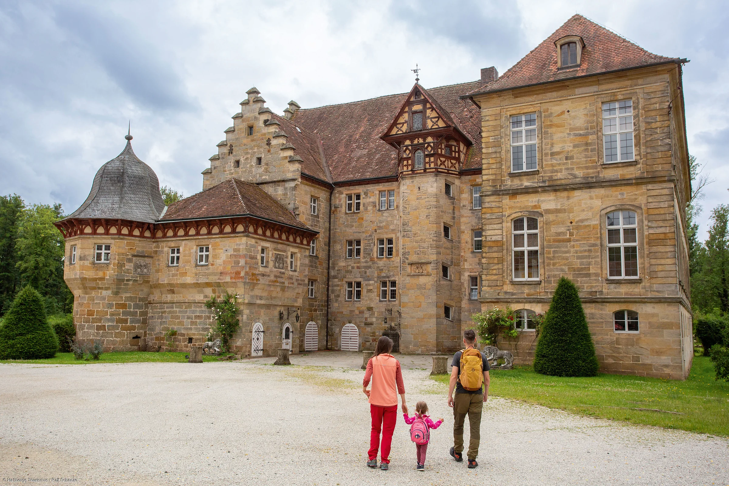 Familie mit Kind und Rucksäcken geht auf ein historisches Schloss mit Sandsteinmauern zu.