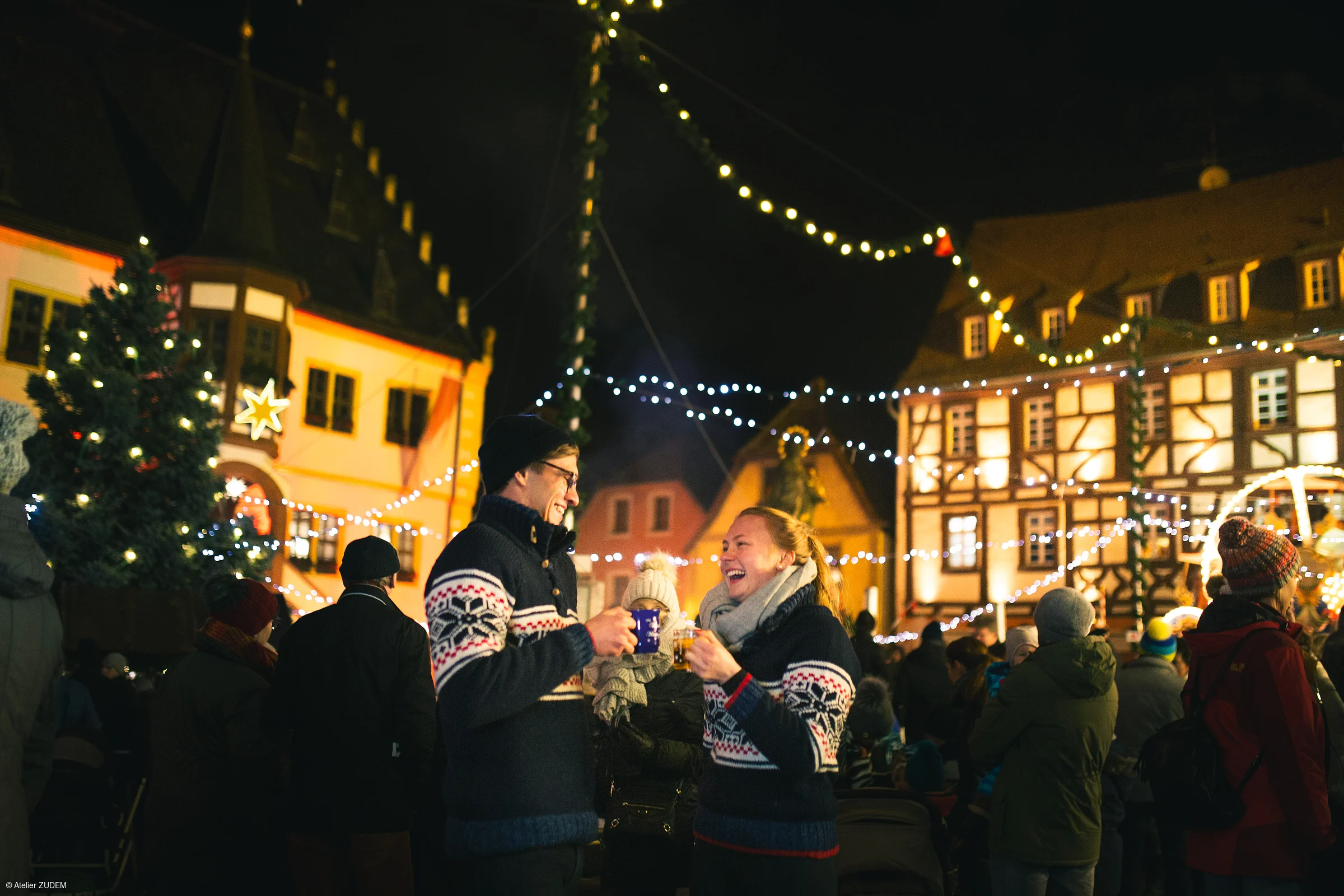 Paar in Winterkleidung trinkt Glühwein auf einem beleuchteten Weihnachtsmarkt bei Nacht.
