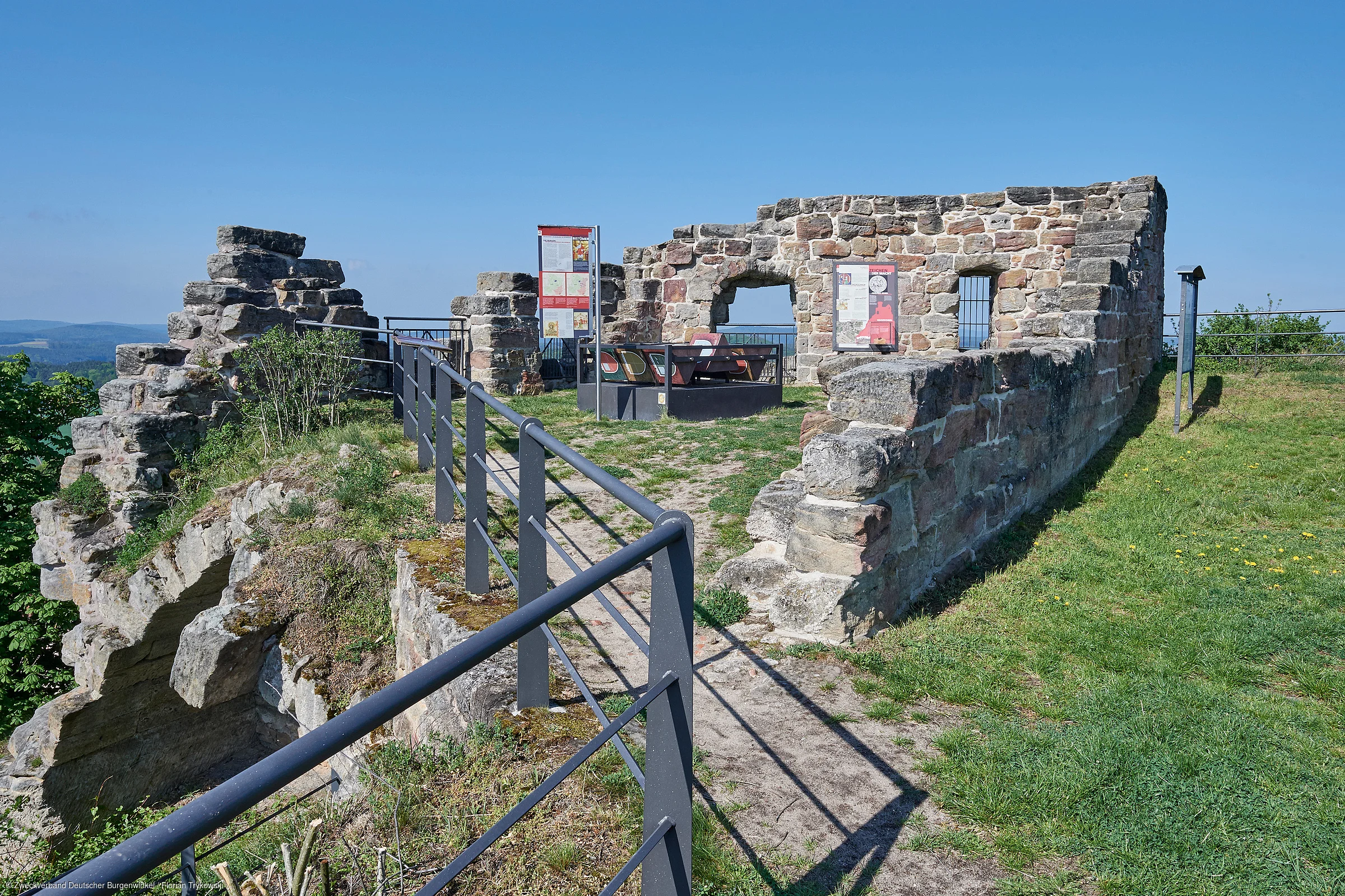 Ruine einer alten Steinmauer mit Infotafeln und Sitzgelegenheiten auf einer Anhöhe bei klarem Himmel.