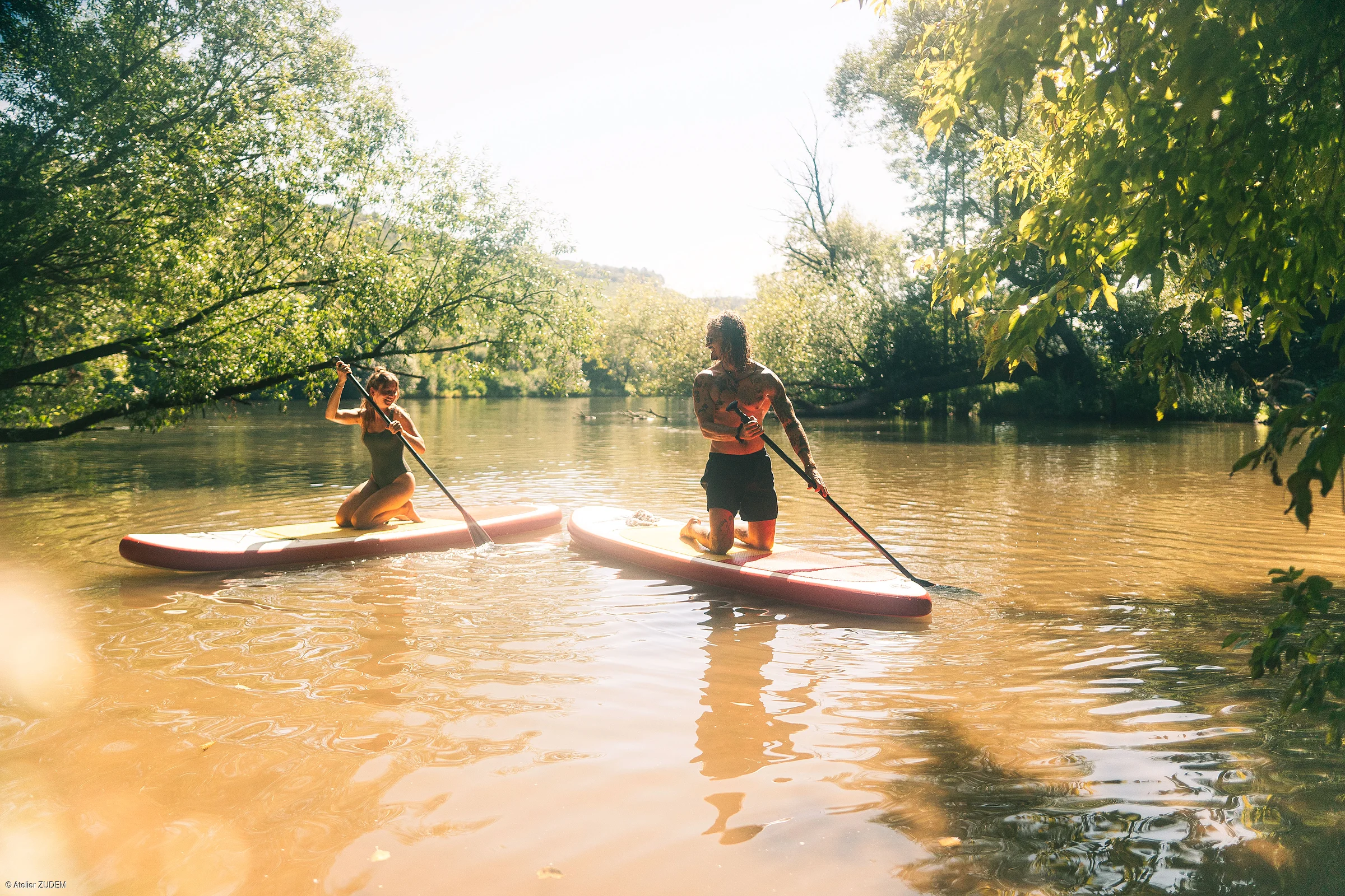 Zwei Personen knien auf Stand-up-Paddle-Boards auf einem Fluss, umgeben von Bäumen im Sonnenlicht.