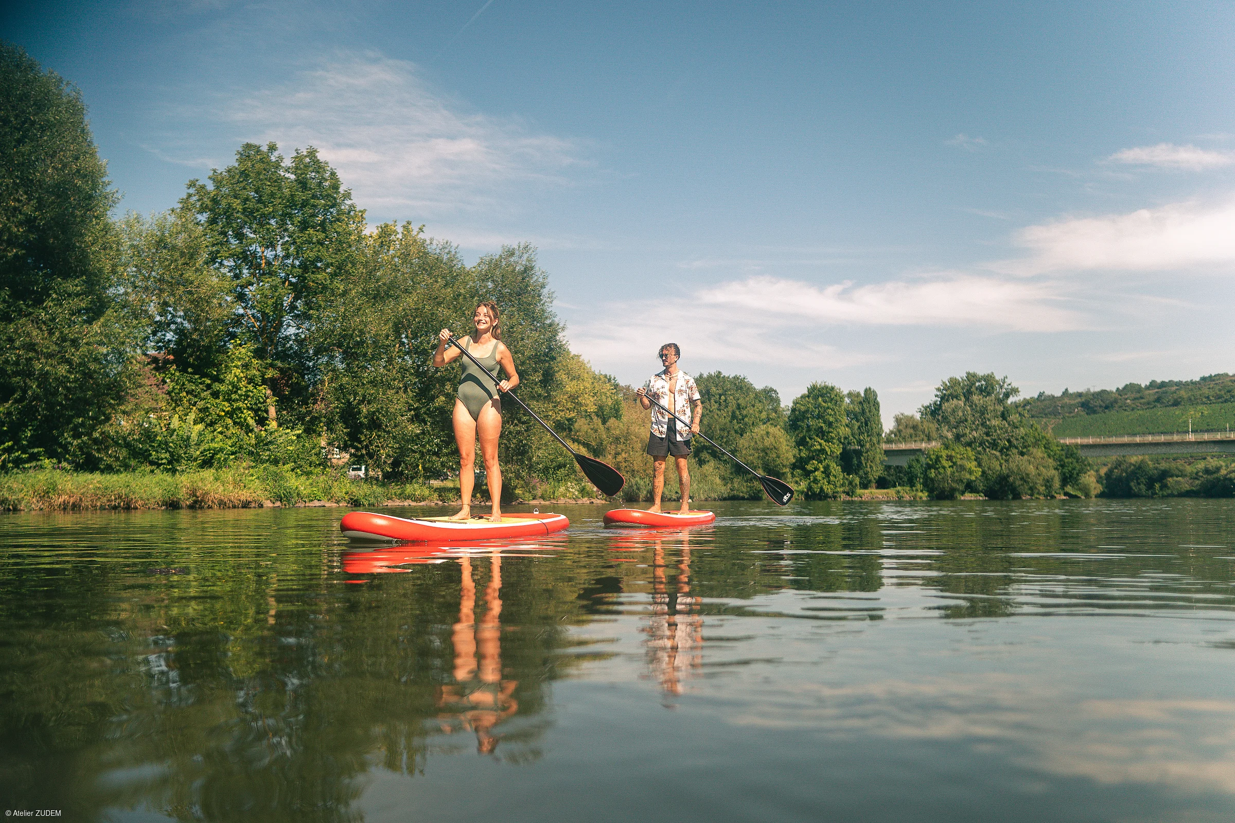 Zwei Personen stehen auf roten Stand-Up-Paddle-Boards auf einem ruhigen Fluss mit Bäumen am Ufer.