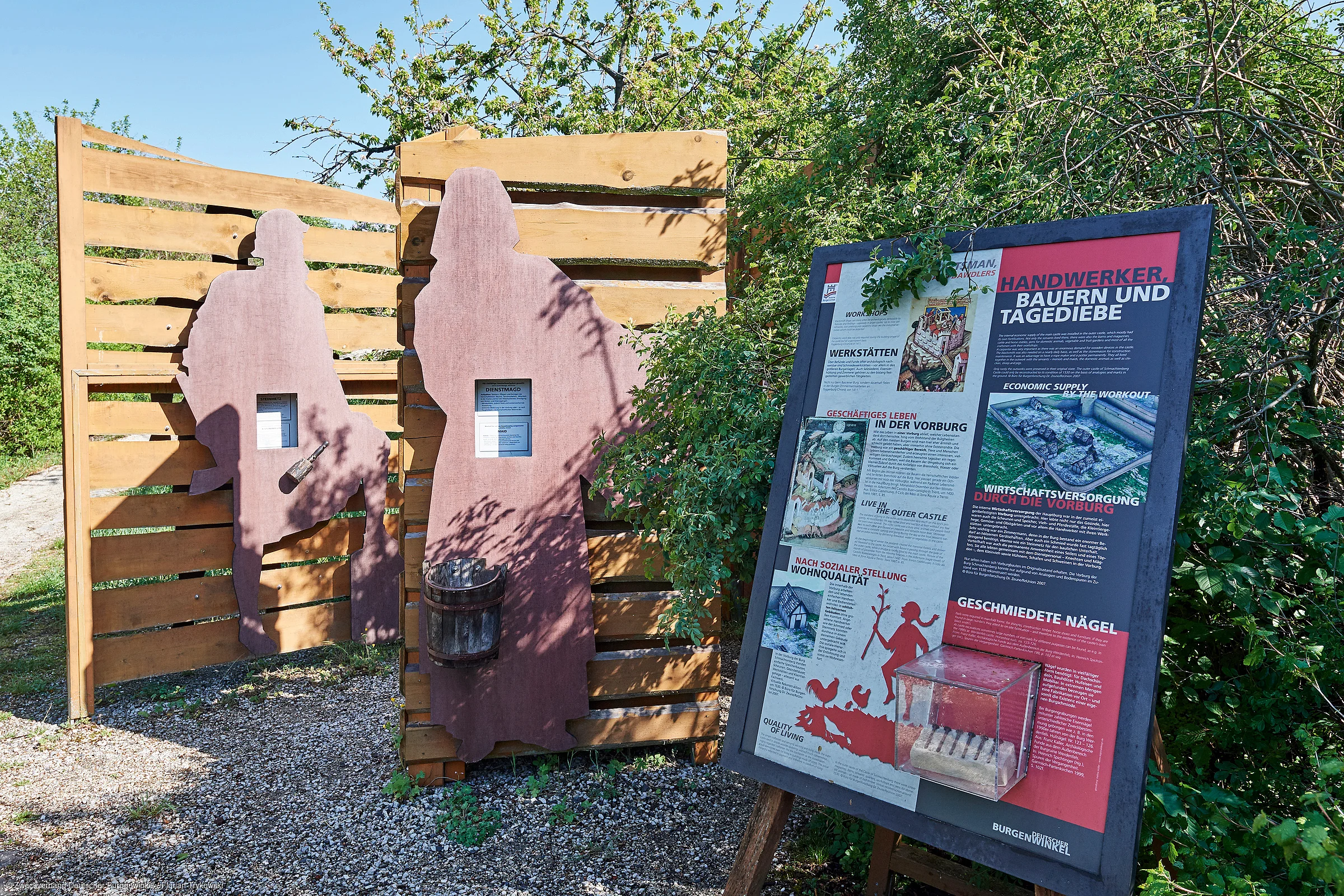 Informationsschild und zwei Holztafeln mit Figuren von Handwerkern im Freien bei Sonnenschein