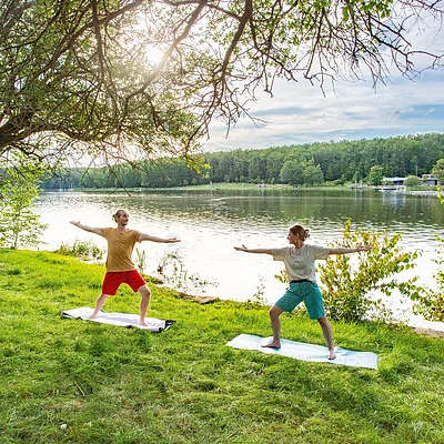 Zwei Personen machen Yoga auf Matten am Ufer eines Sees, umgeben von Bäumen und Gras.