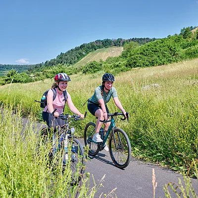 Zwei Frauen fahren mit Fahrrädern auf einem schmalen Weg durch eine grüne Wiesenlandschaft bei klarem Himmel.