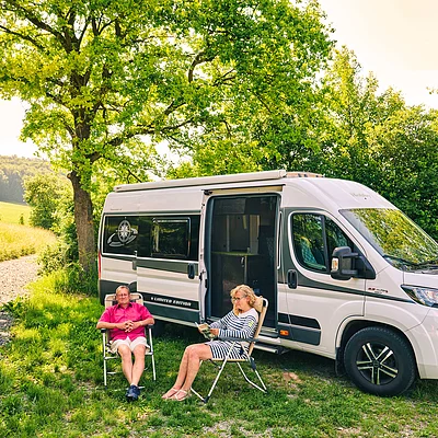 Zwei Personen sitzen auf Campingstühlen neben einem weißen Wohnmobil auf einer Wiese unter Bäumen.