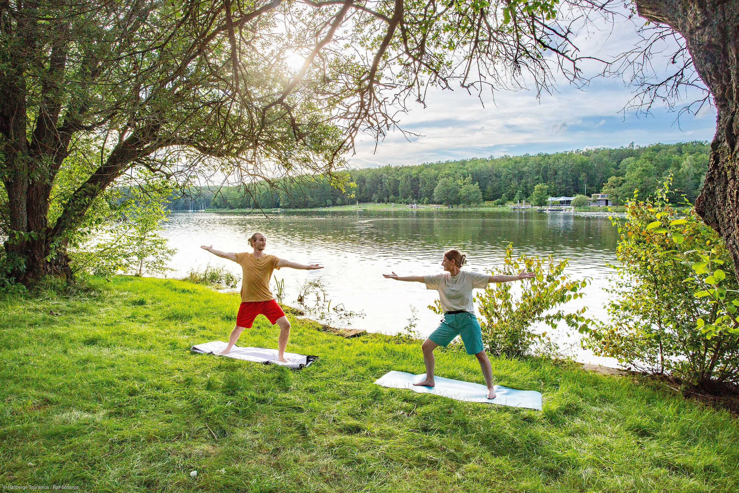 Zwei Personen machen Yoga auf Matten am Ufer eines Sees, umgeben von Bäumen und Gras.