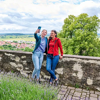 Zwei Frauen machen an einer Steinmauer mit Blick auf Landschaft und Dorf ein Selfie bei bewölktem Himmel.