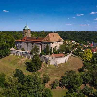 Luftaufnahme einer Burg auf einem bewaldeten Hügel mit Dorf im Hintergrund unter blauem Himmel.
