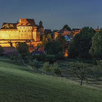 Beleuchtete Burg auf einem Hügel mit Bäumen und Wiesen im Vordergrund bei Abenddämmerung
