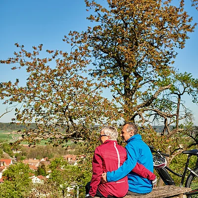Älteres Paar sitzt auf Holzbank im Grünen, Fahrrad und Rucksack stehen daneben, Dorf im Hintergrund bei klarem Himmel