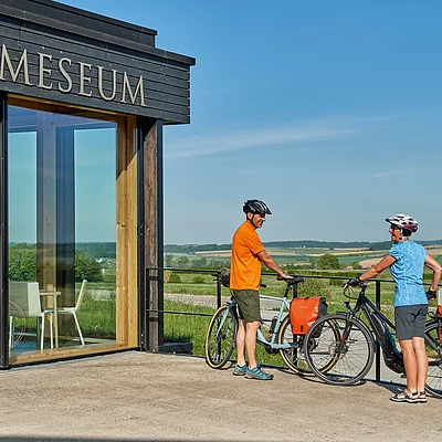 Zwei Personen mit Fahrrädern stehen vor dem Gebäude des Limeseums bei klarem Himmel und grüner Landschaft.