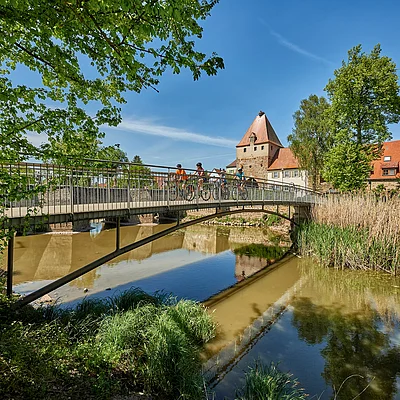 Brücke über Fluss mit Radfahrern, Bäumen und historischen Gebäuden bei klarem Himmel im Hintergrund.