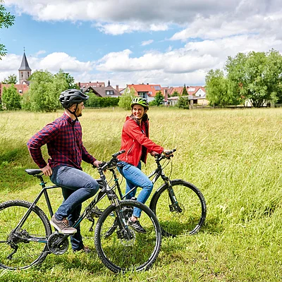 Zwei Personen mit Fahrradhelmen auf Mountainbikes auf einer Wiese vor einem Dorf mit Häusern und Kirche im Hintergrund.
