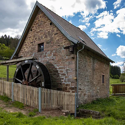 Altes Steinhaus mit Wasserrad und Holzzaun in grüner Landschaft unter bewölktem Himmel.
