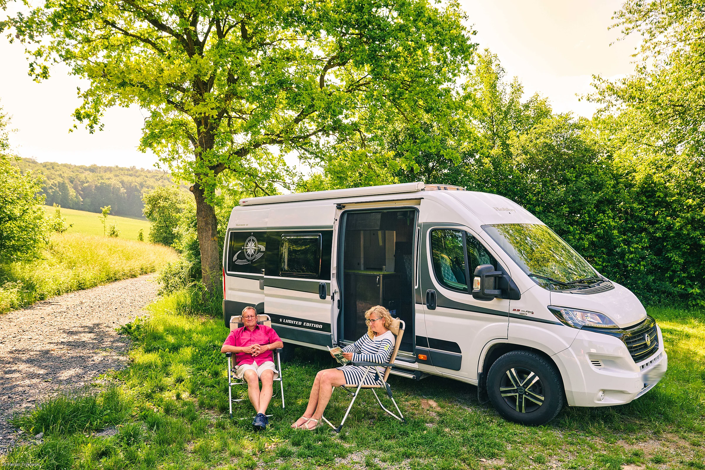 Zwei Personen sitzen auf Campingstühlen neben einem weißen Wohnmobil auf einer Wiese unter Bäumen.