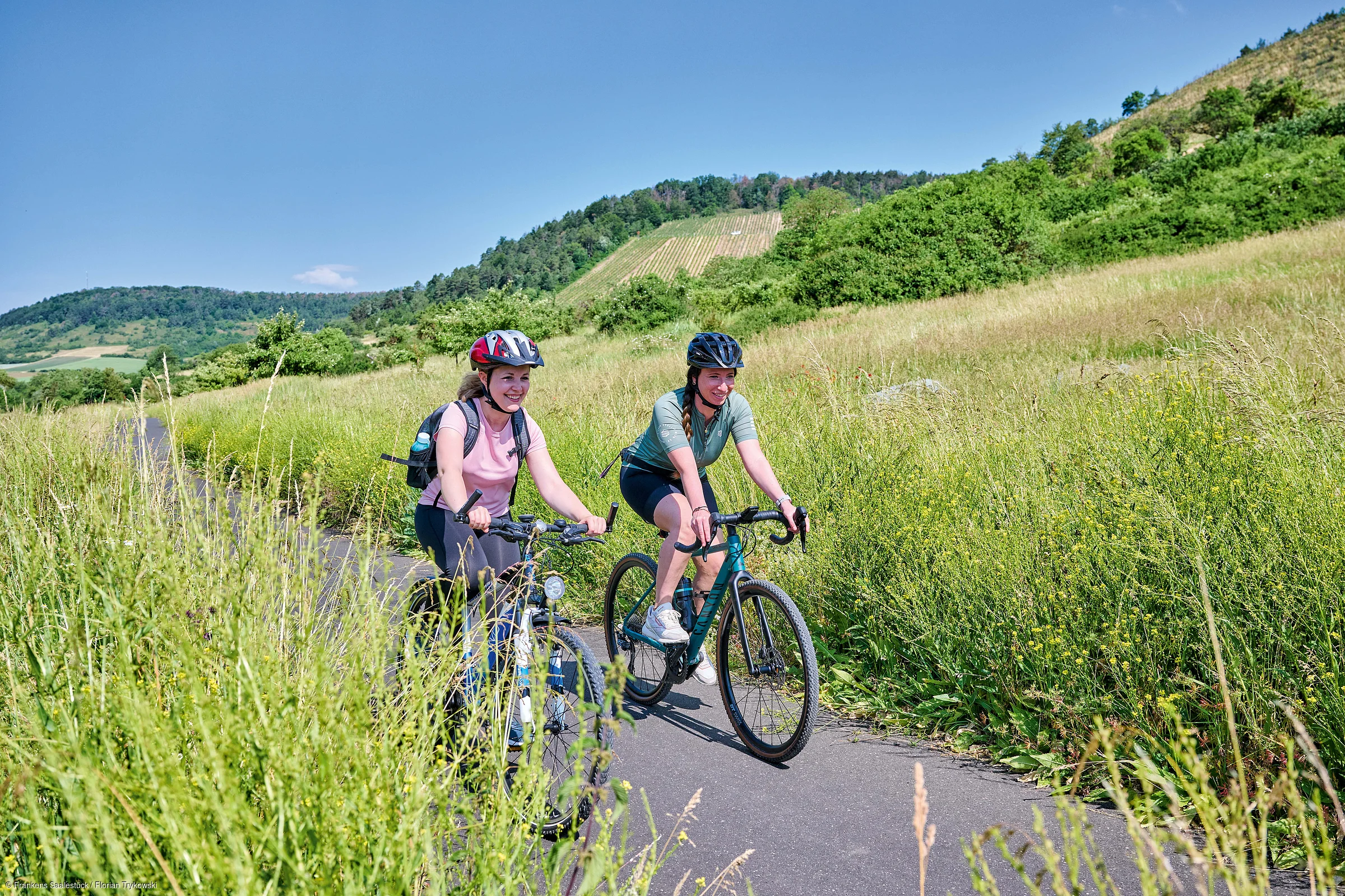 Zwei Frauen fahren mit Fahrrädern auf einem schmalen Weg durch eine grüne Wiesenlandschaft bei klarem Himmel.