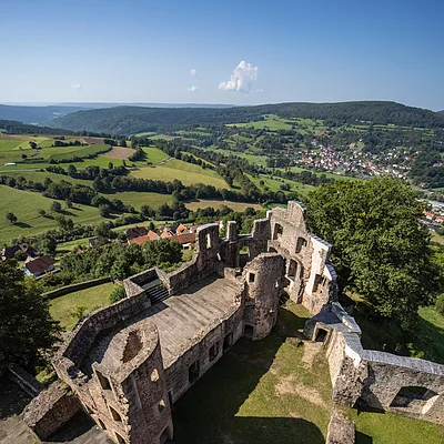 Blick von oben auf die Ruine einer Burg mit umliegender grüner Landschaft und Dorf im Hintergrund.