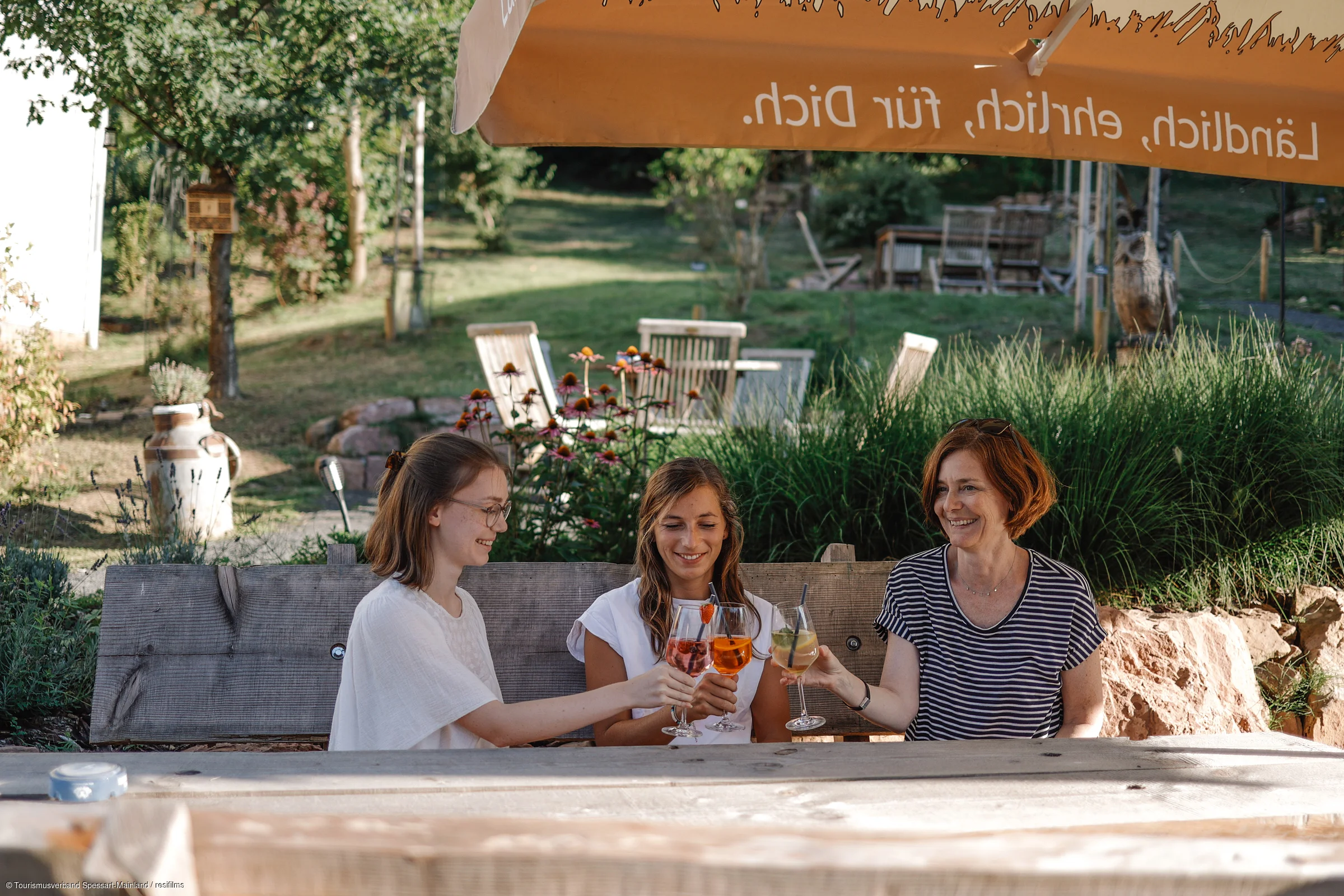 Drei Frauen sitzen draußen an einem Holztisch und stoßen mit Getränken unter einem Sonnenschirm an.