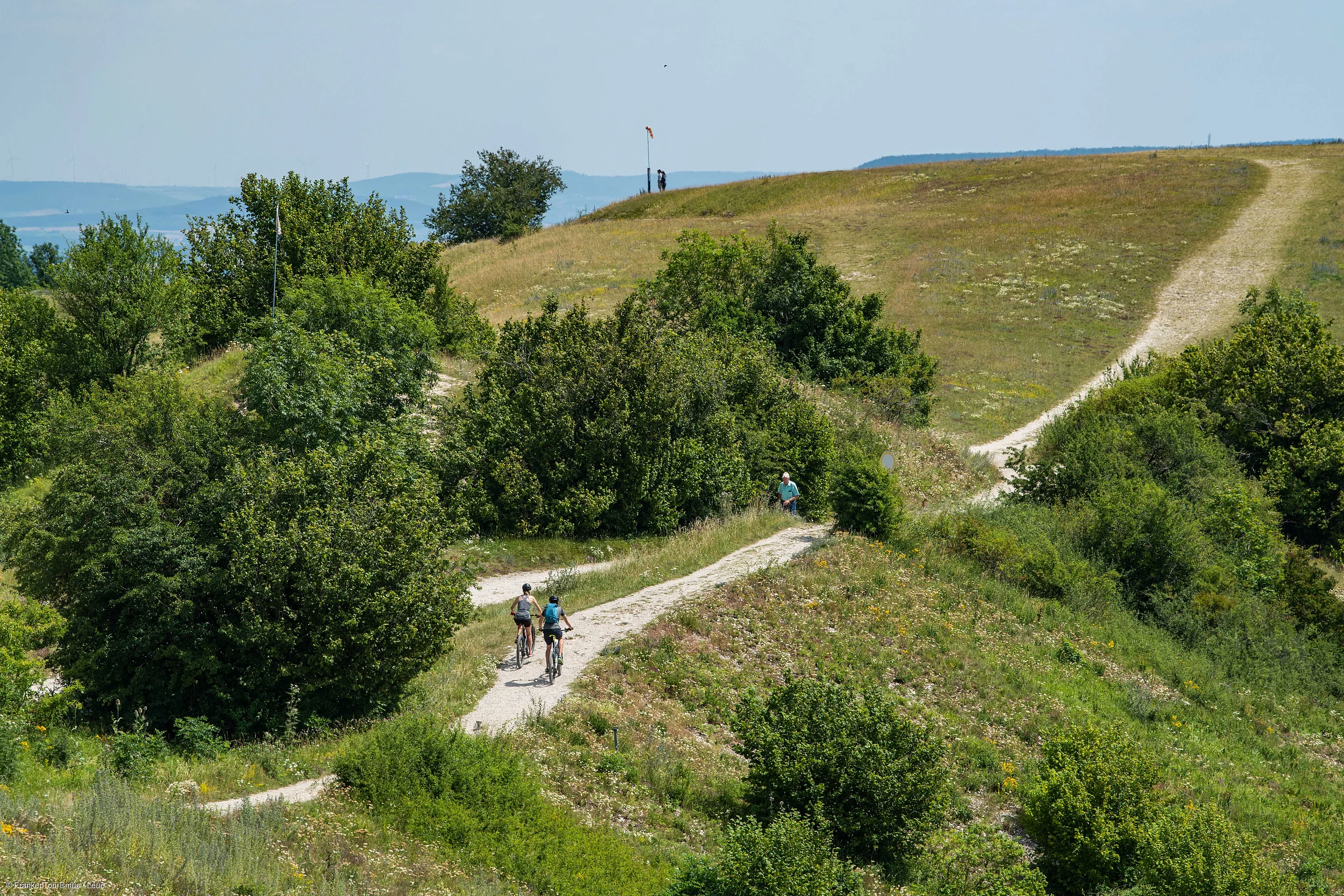 Zwei Radfahrer auf einem geschwungenen Weg durch grüne Hügel mit Büschen und Bäumen.