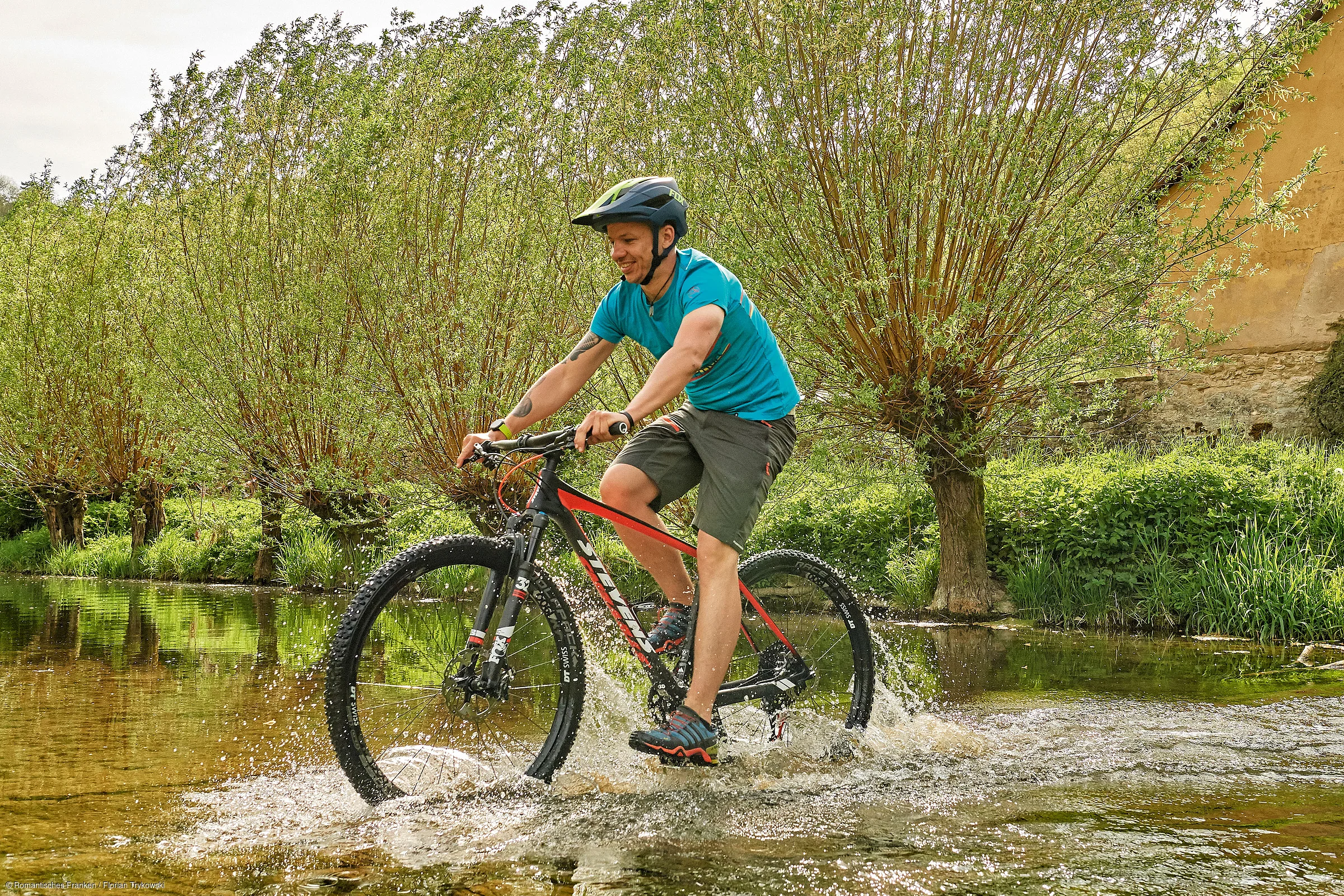 Mann mit Helm fährt Mountainbike durch flaches Wasser vor grünen Büschen und altem Gebäude.