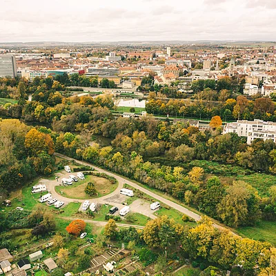 Luftaufnahme einer Stadt mit Herbstbäumen, Wohnmobilstellplatz und Fluss im Vordergrund.