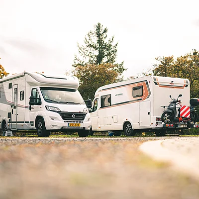 Zwei weiße Wohnmobile stehen auf einem Parkplatz vor herbstlich gefärbten Bäumen, eines mit montiertem Motorroller.