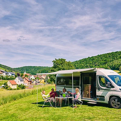Vier Personen sitzen an einem Tisch unter einer Markise neben einem weißen Wohnmobil auf einer Wiese bei Dorf und Wald