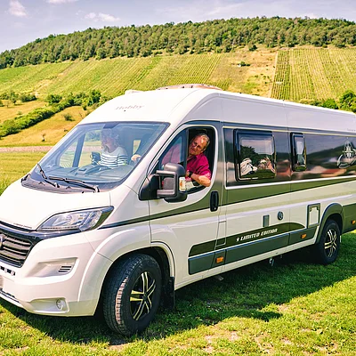 Weißer Wohnmobil-Van mit zwei Personen auf einer grünen Wiese vor bewaldeten Hügeln und Weinbergen.
