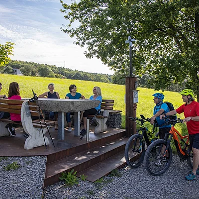 Sechs Frauen sitzen an einem runden Tisch auf einer Bank, zwei Männer mit Fahrrädern stehen daneben unter einem Baum.