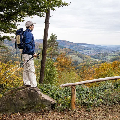 Wanderer mit Rucksack und Stöcken steht auf Felsen und blickt auf herbstliche Landschaft mit Hügeln und Tal.
