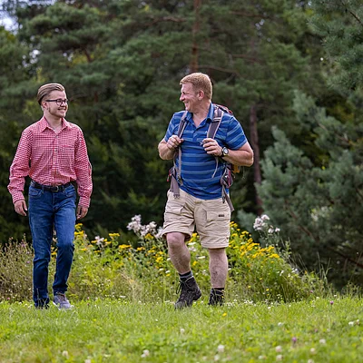 Zwei Männer wandern auf einem Wiesenweg, umgeben von Bäumen und Wildblumen.