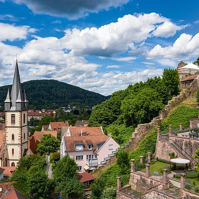 Blick auf eine Kirche mit spitzem Turm, Häuser, grüne Hügel und eine alte Burgmauer mit Turm und Flagge.