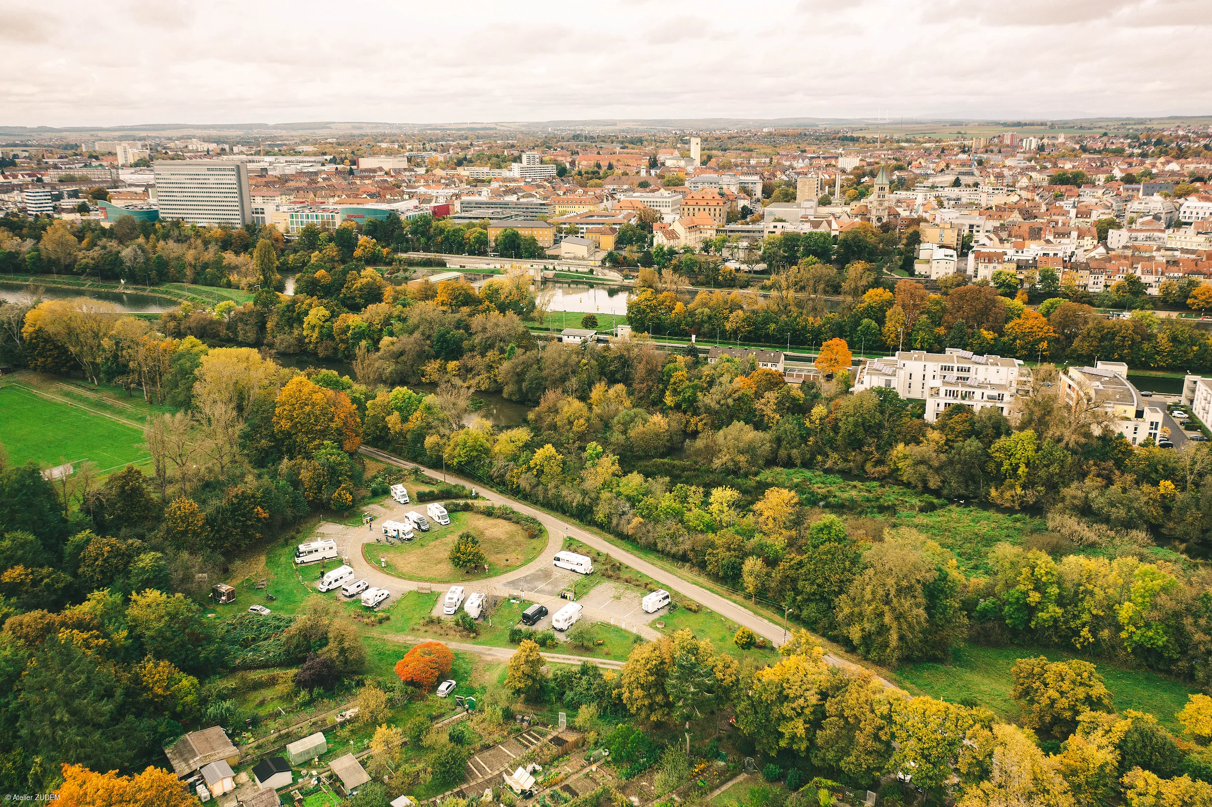 Luftaufnahme einer Stadt mit Herbstbäumen, Wohnmobilstellplatz und Fluss im Vordergrund.