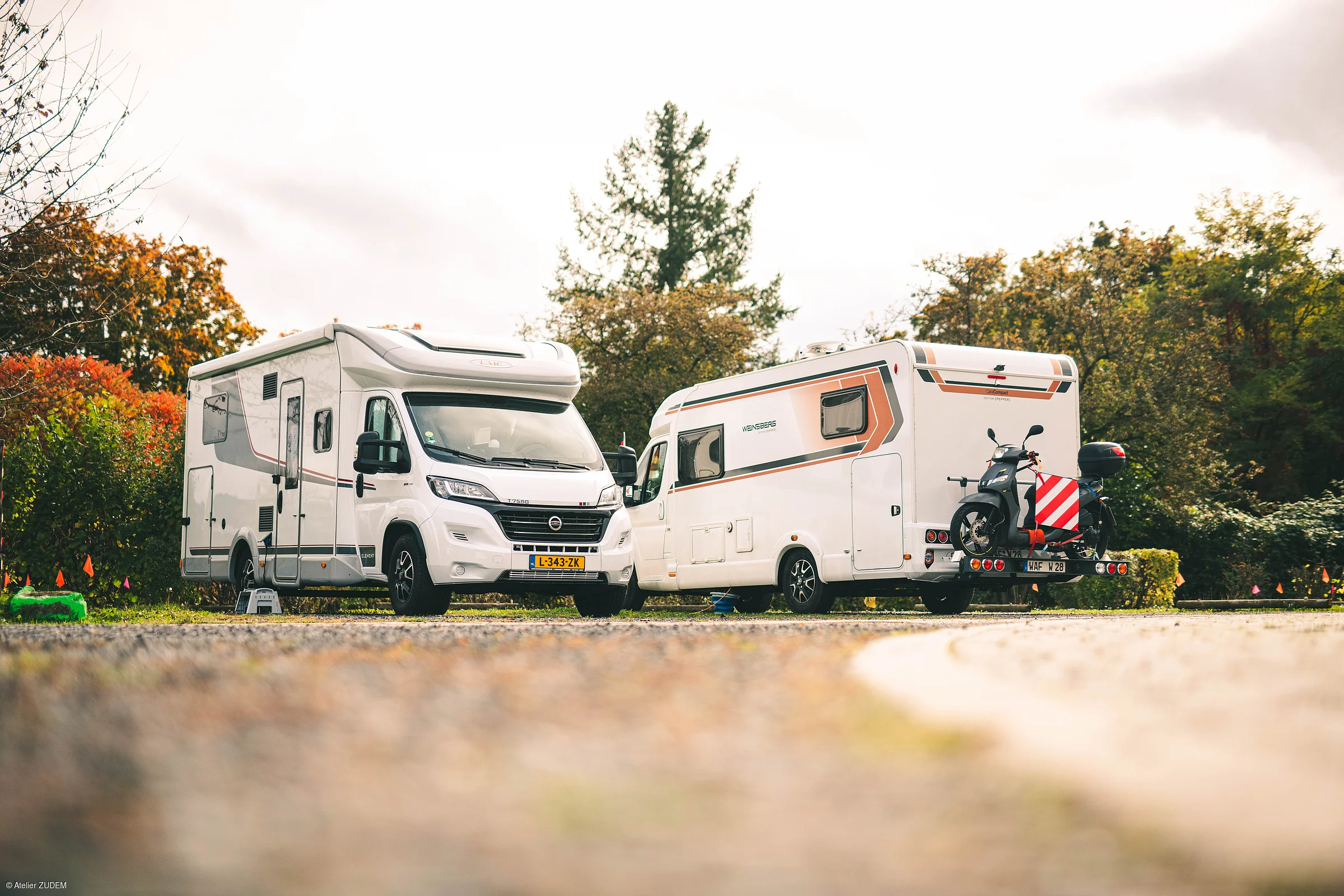 Zwei weiße Wohnmobile stehen auf einem Parkplatz vor herbstlich gefärbten Bäumen, eines mit montiertem Motorroller.