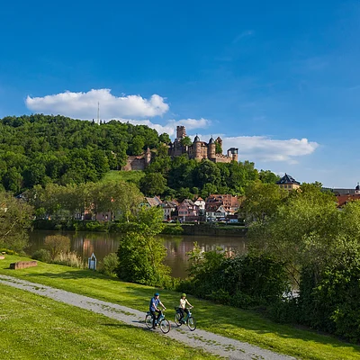 Zwei Radfahrer auf Weg am Fluss mit Schloss auf bewaldetem Hügel und Dorf im Hintergrund bei blauem Himmel.