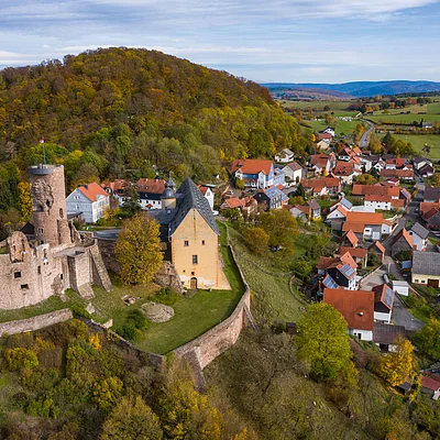 Luftaufnahme einer Burganlage mit umliegendem Dorf und herbstlich gefärbtem Wald auf einem Hügel.