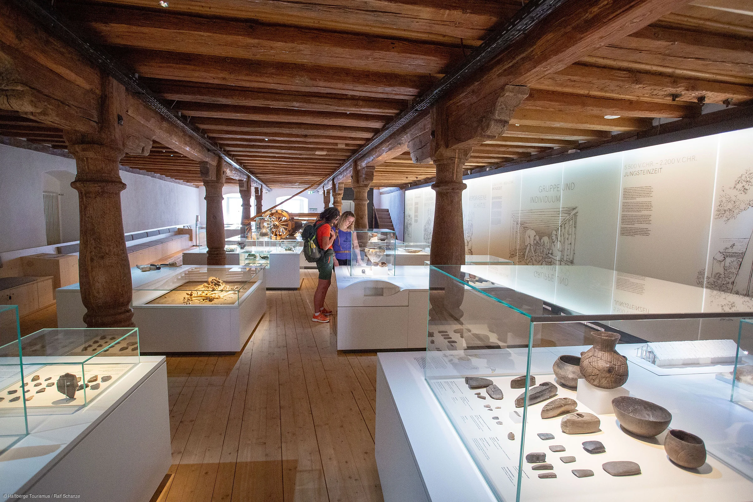 Museumsausstellung mit historischen Artefakten in Vitrinen und zwei Personen, die diese betrachten, Holzbalkendecke und Holzboden