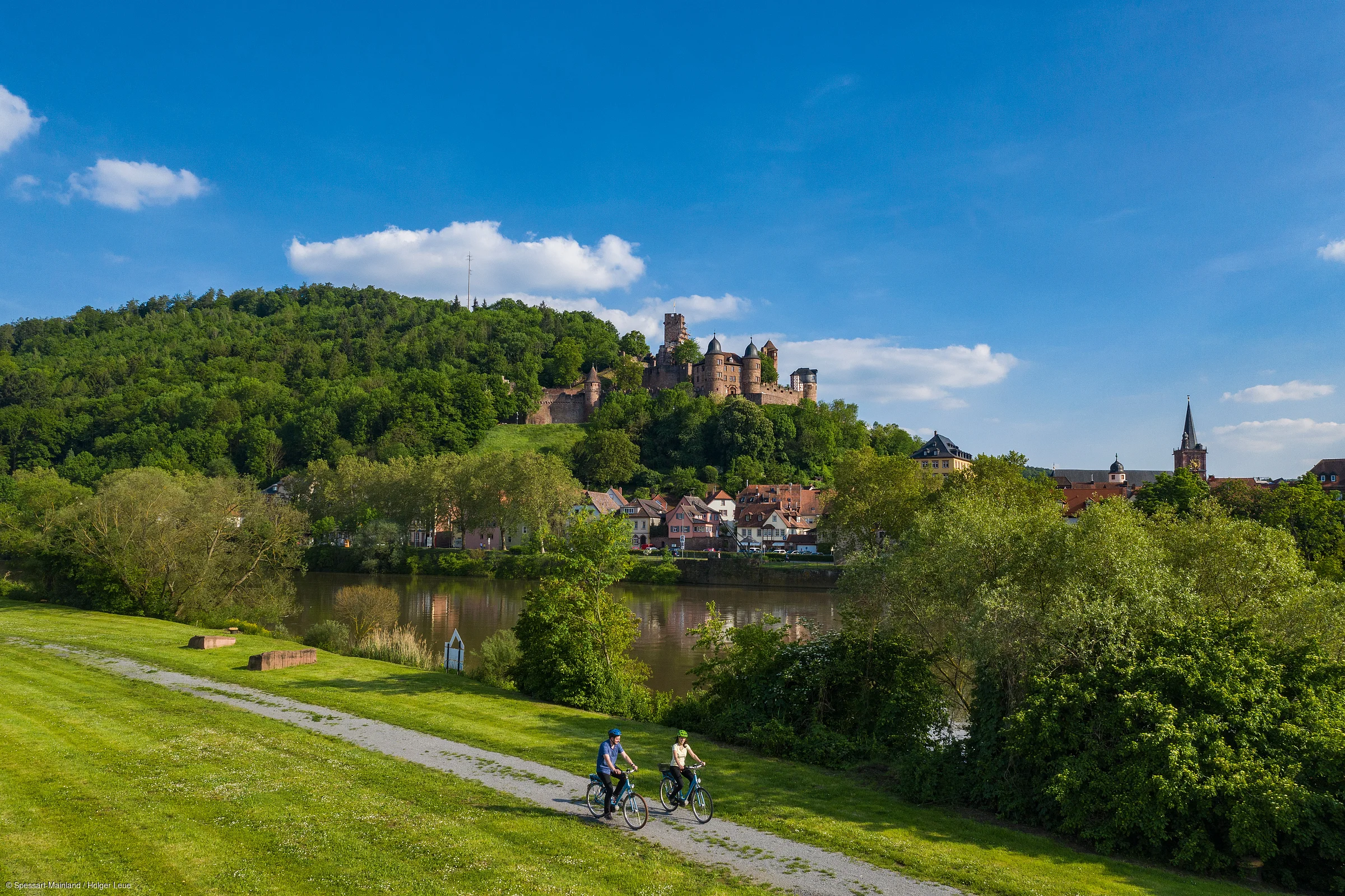 Zwei Radfahrer auf Weg am Fluss mit Schloss auf bewaldetem Hügel und Dorf im Hintergrund bei blauem Himmel.