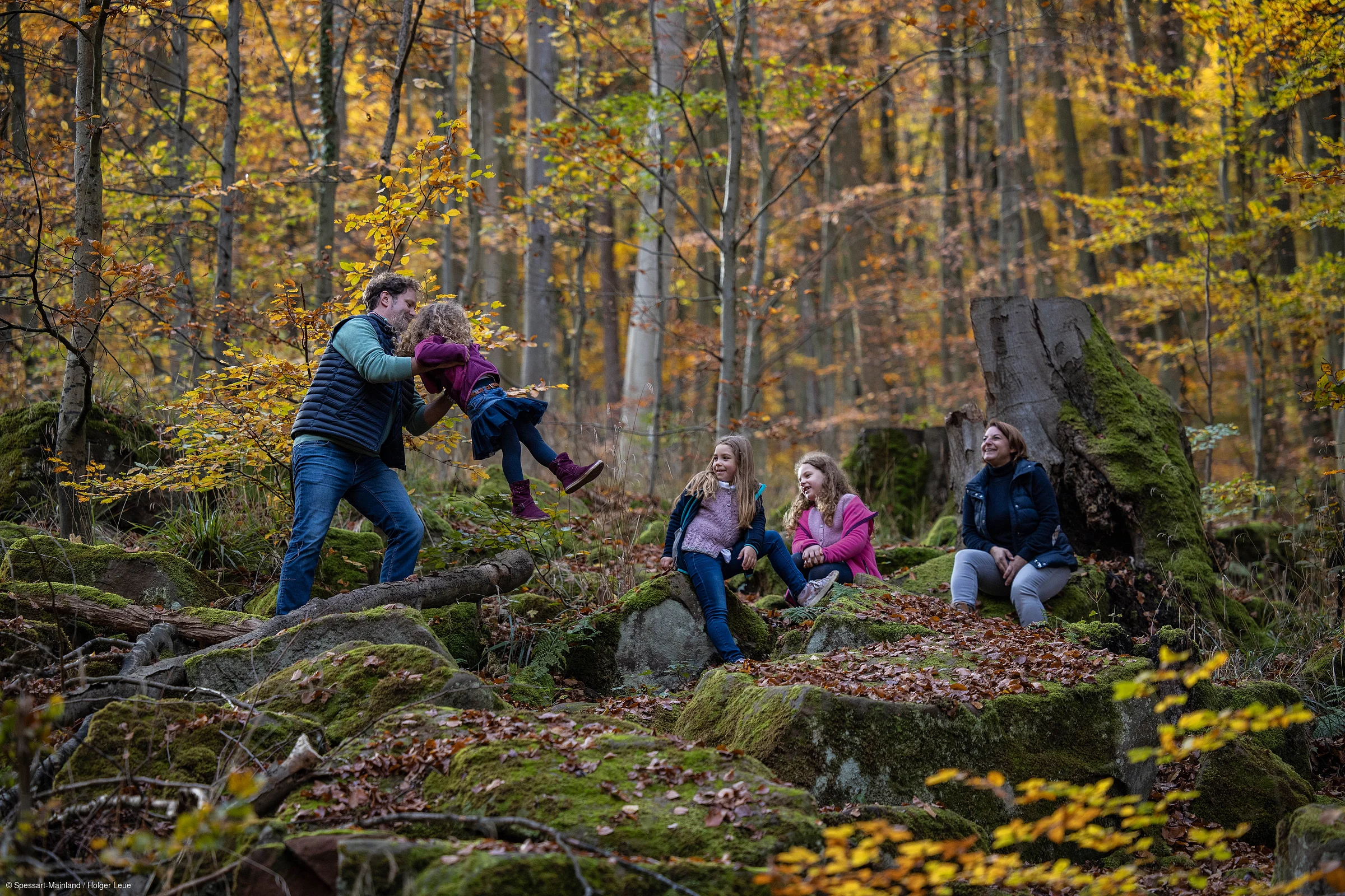 Vater spielt mit Kind, zwei Mädchen und Frau sitzen auf moosbedeckten Steinen im herbstlichen Wald.